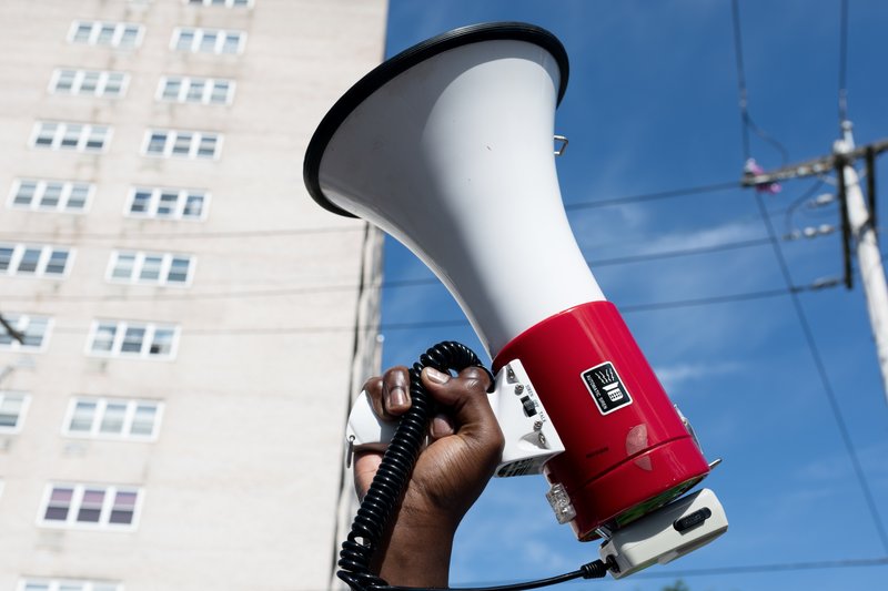 Image of a hand holding up a red and white bullhorn with a tall tan building in the background